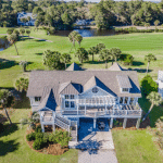 A large house with a wraparound porch overlooks a green golf course and a pond, surrounded by trees and palm trees.
