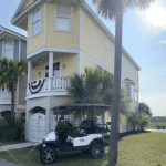 A white golf cart is parked in front of a yellow beach house decorated with a patriotic banner.