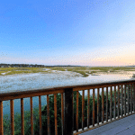 Wooden deck overlooks a marsh with water and grass under a clear blue sky at sunset.