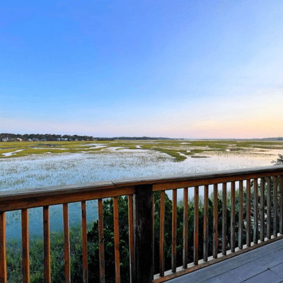 Wooden deck overlooks a marsh with water and grass under a clear blue sky at sunset.