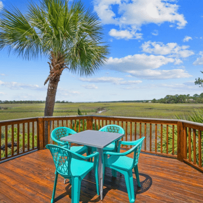 A wooden deck with four teal chairs, a table, a palm tree, and a scenic marsh view under a blue sky.