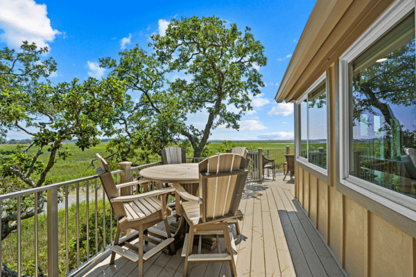 Wood deck with wooden chairs and table, surrounded by trees, overlooking a green landscape under a blue sky.