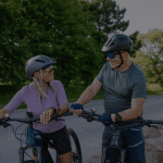 Two adults wearing helmets smile at each other while standing with bicycles on a rural road.