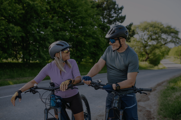 Two adults wearing helmets smile at each other while standing with bicycles on a rural road.