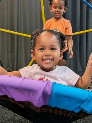 Two smiling children play; one sits on a colorful swing, the other stands behind her. Perfect for a rainy Fripp day or anytime!