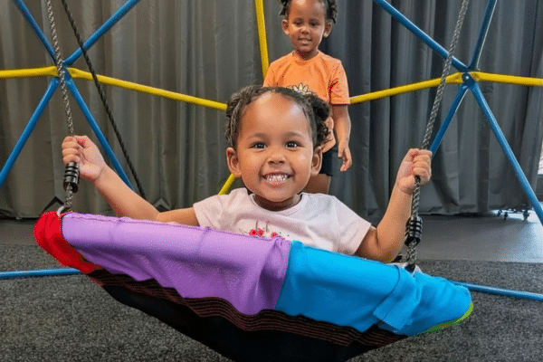 Two smiling children play; one sits on a colorful swing, the other stands behind her. Perfect for a rainy Fripp day or anytime!
