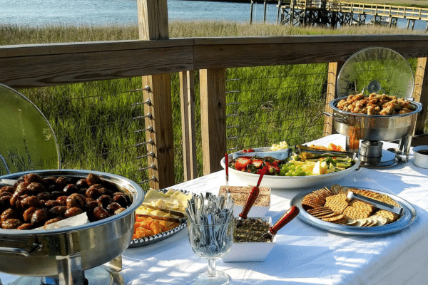 Buffet table with various appetizers and snacks set outdoors near water and a wooden dock.