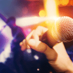 Hand holding a microphone on stage with blurred musicians and colorful lights in the background.