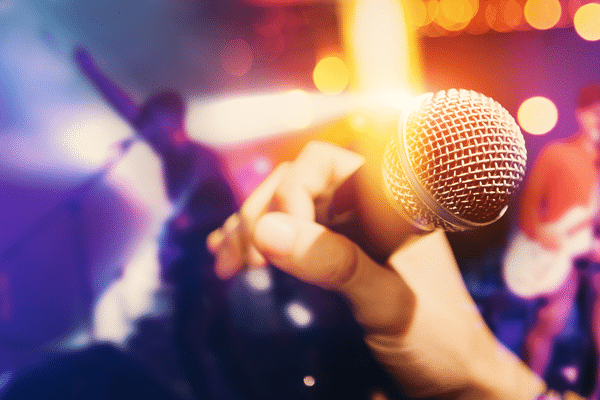 Hand holding a microphone on stage with blurred musicians and colorful lights in the background.