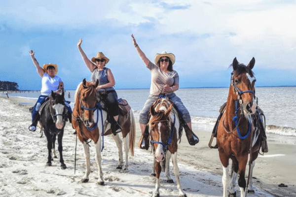 Four people on horses by the ocean, wearing hats and waving on a sandy beach under a cloudy sky.