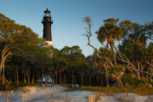 A black-and-white lighthouse rises above palm trees and sand dunes at sunset.