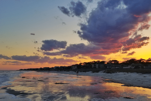 Colorful sunset sky with clouds over a beach, houses, and calm water reflecting the light.