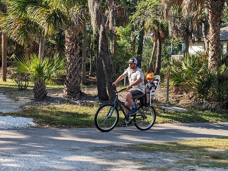 Man rides a bike with a child in a rear seat, surrounded by palm trees on a sunny day on the Fripp Island bike trail.