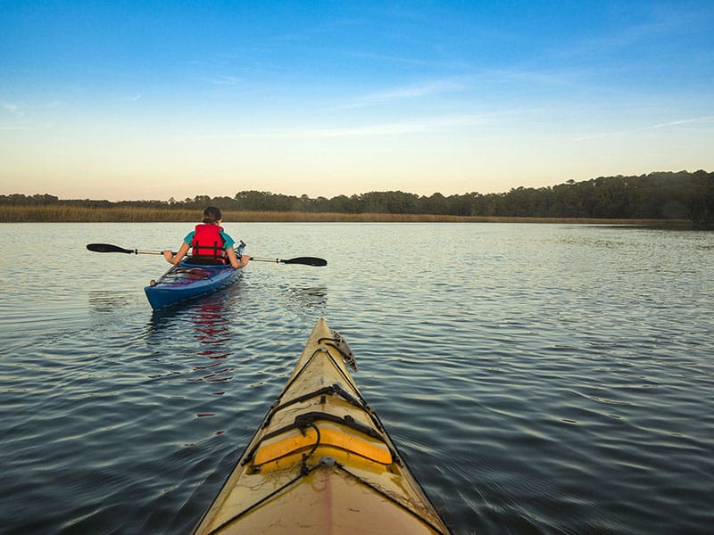 A person kayaks on the waterways surrounding Fripp Island at sunset, seen from another kayak following behind.