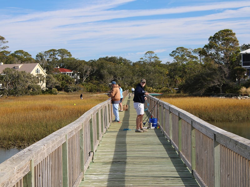 Two people are fishing on Wardles Landing surrounded by marsh grass under a partly cloudy sky.