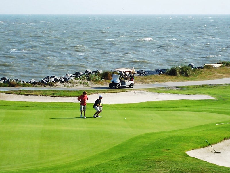 Two people on Ocean Point green near the ocean, with a golf cart and waves in the background. Try your luck at this stunning links course.