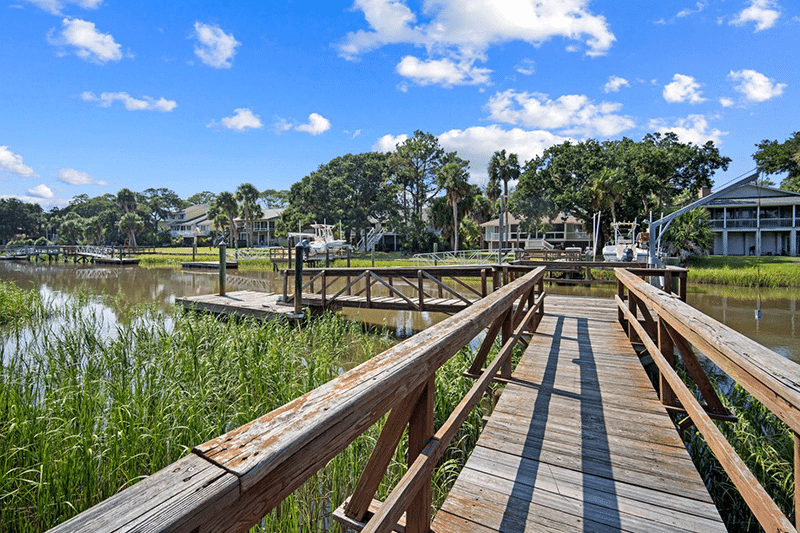 Wooden dock over marsh grass leading to water, with houses and trees in the background under a blue sky.
