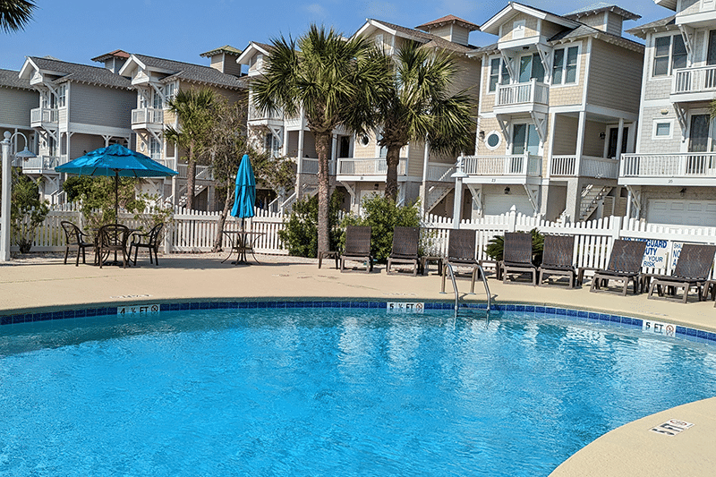 Blue pool with lounge chairs and umbrellas, palm trees, and beach houses in the background under a clear sky.