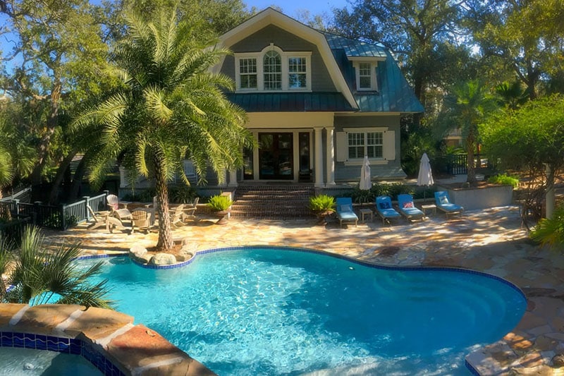 A house with a large pool, palm trees, lounge chairs, and umbrellas in a sunny backyard.