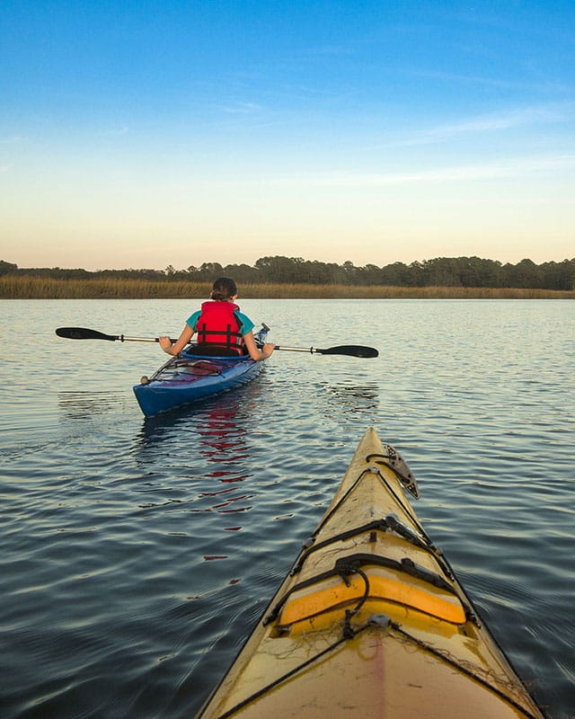 Two kayakers paddle on a calm lake at sunset, one ahead in a blue kayak, trees line the distant shore.