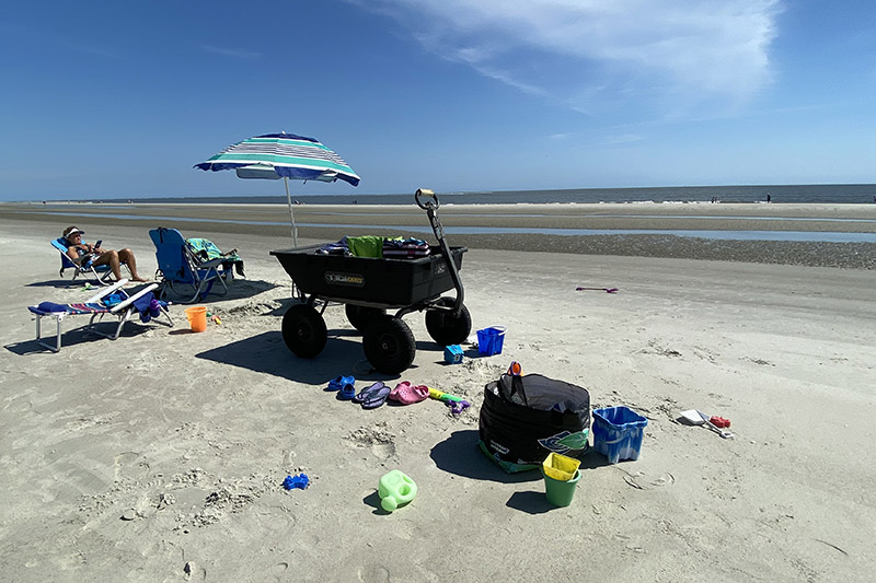 Beach scene with chairs, umbrella, wagon, and sand toys on a sunny day near the ocean.