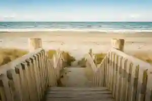 Wooden stairs lead down to a sandy beach with waves and a clear blue sky in the background.
