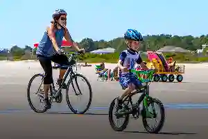 Woman and child wearing helmets ride bicycles on a sunny day near people sitting on a beach.
