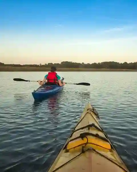 Two people kayaking on calm water at sunset, one in a blue kayak ahead of the photographer.