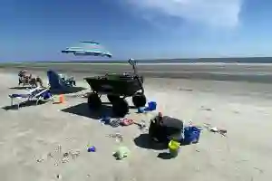 A cart, beach chairs, toys, and a striped umbrella are set up on a sandy beach with a blue sky overhead.
