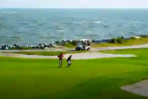 Two people on a grassy golf course near the ocean, with a golf cart parked on a path in the background.