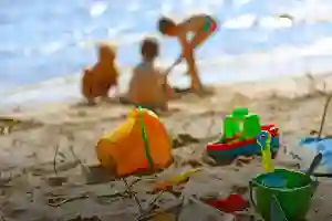Children play on a sandy beach; colorful toys and buckets are scattered in the foreground.